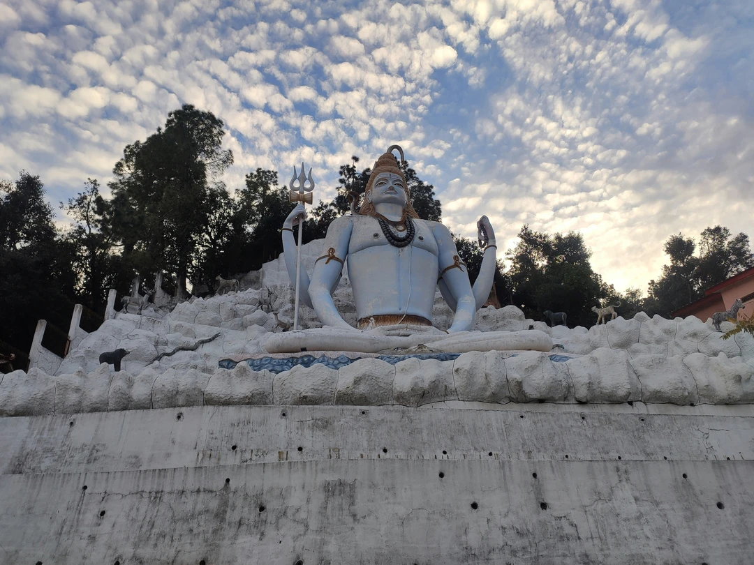 The large, pale blue statue of a meditating Lord Shiva with his trident, set against a dramatic, cloud-filled sky in Ranikhet