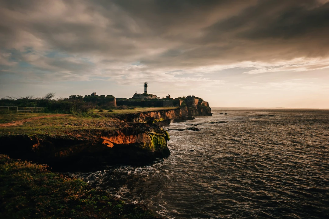 The historic Diu Fort and its lighthouse stand on a rocky cliff overlooking the Arabian Sea under a moody, golden sunset sky