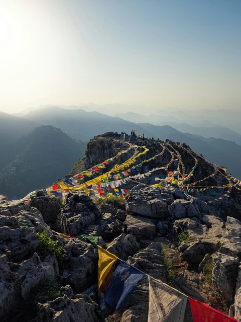 The rocky summit of George Everest Peak adorned with colorful Tibetan prayer flags, overlooking layers of hazy Himalayan hills